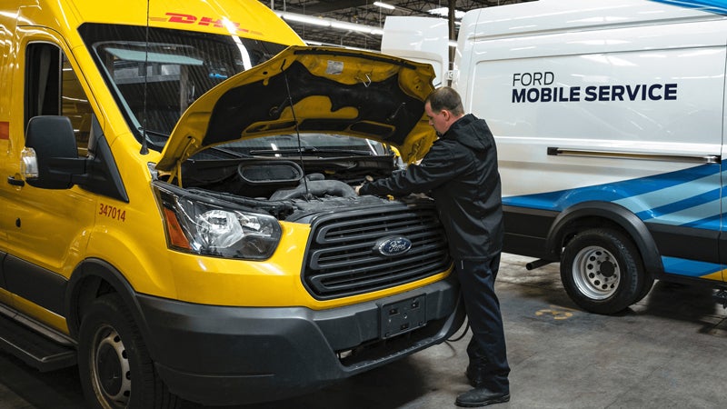 A technician repairs a yellow DHL delivery van, with its hood up, next to a white Ford Mobile Service van.